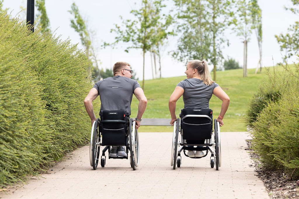 Eine junge Frau und ein junger Mann sitzen jeweils in einem Küschall Aktivrollstuhl und fahren nebeneinander her. Sie sind von hinten zu sehen.