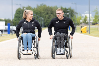 Eine junge Frau und ein junger Mann sitzen jeweils in einem Küschall Aktivrollstuhl und fahren nebeneinander her.