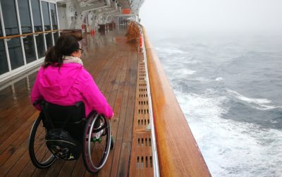 Eine Frau sitzt in ihrem Rollstuhl. Sie ist von hinten zu sehen. Sie befindet sich auf einem Schiff und schaut auf das Meer.
