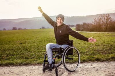 Ein junger Mann im schwarzen Pulli sitzt in einem Aktivrollstuhl der Küschall K-Serie. Er lacht und hat die Arme ausgestreckt.