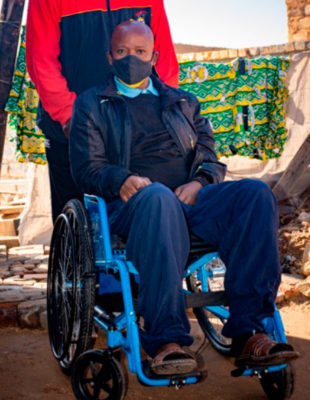 Ein Mann sitzt im Rollstuhl und wird von einer anderen Person geschoben. Er trägt eine Maske.